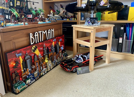 A photo showing the Lego Gotham City Skyline set positioned on the floor in front of a wooden trunk. The set has lights below police blimps, enhancing their spot lights, as well as a lot search light on the roof of the Police Station, and more lights lighting up the Batsignal in the sky. In front of it is the Lego 1960’s Batmobile set, and above that the Lego set for the 1989 Batwing.
