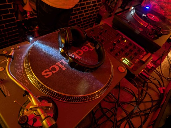 photo of a turntable in the front, visually set in a club setting, with a 3 channel mixer and a second turntable in the background. lighting is cozy reddish. there's a dj headphones on the turntable in the front, but no vinyl.