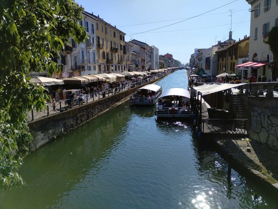 Das Bild zeigt einen Kanal in einer italienischen Stadt, wahrscheinlich Venedig oder eine ähnliche Stadt mit Kanälen. An den Ufern stehen Gebäude im venezianischen Stil, mit Arkaden und Balkonen. Viele Markisen und Sonnenschirme bedecken die angrenzenden Geschäfte und Cafés. Der Kanal ist belebt; viele Menschen spazieren entlang der Uferpromenade und zwei Ausflugsboote fahren auf dem Wasser. Die Atmosphäre ist sonnig und lebhaft, mit einer typisch italienischen Urlaubsstimmung.