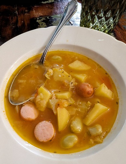 Dunkler Tisch, weißer Suppenteller mit rotgelber Gemüsesuppe darin. Es sind Kartoffelstücke, Kohl, dicke Bohnen und Würstchenscheiben erkennbar.  Ein silberner Löffel liegt im Teller.