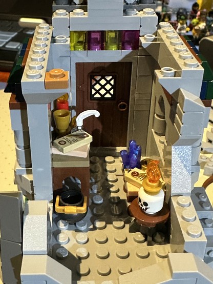 Bags 22 & 23 of LEGO set 21348. Interior of arcane workshop on first floor of the tower. Work surfaces hold spell scrolls, writing utensils, candles, and a crystal. A cauldron sits on the floor and a wooden stool holds a candle set atop a human skull. Creepy!