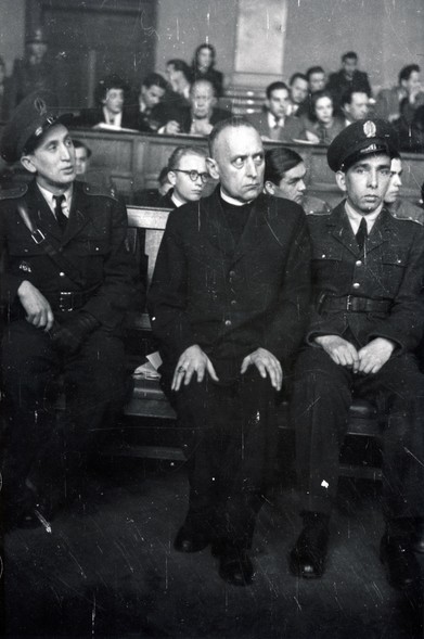 A monograph photo of a man in a dark clerical suit sitting on a courtroom wooden bench with two men in military-style uniforms sitting on either side of him. The man has a severe look on his face and is looking to his left, as is one of his guards. The other guard is looking directly at the camera. In the background are various people also sitting on wooden benches in the courtroom.