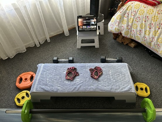 A set of coloured weights plus bar in front of a bench with a blue towel on it. Behind the bench is another set of 2 weights, a small step stool with my iPad set to the class I’ll be doing. A single bed with a light coloured doona is on the right with net curtains visible in the back left.