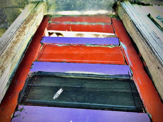 An old, rickety door stands tall in a wooden doorway, with red as its primary color, purple as its secondary, and one of two central slats painted a deep jungle/aquamarine green as its tertiary, with the other slat missing entirely.