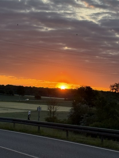 Ein stimmungsvoller Sonnenaufgang über einer ländlichen Landschaft. Die Sonne erhebt sich am Horizont hinter einer Baumreihe und taucht den Himmel in leuchtendes Orange und Rot, während darüber dunkle Wolken dramatisch den Himmel durchziehen. Im Vordergrund führt eine Landstraße mit Leitplanke durch sanfte Felder, auf denen sich der neue Tag langsam entfaltet. Ein Auto ist in der Ferne unterwegs - Symbol für den beginnenden Alltag im Licht eines friedlichen Morgens.