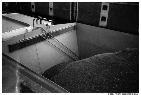 Black and white photograph looking down into the cargo of a river barge in a lock. The cargo is crushed gravel that does not completely fill to hold and there is a ladder dangling from the middle of the ship to the starboard side diagonally inside the hold meeting at the side of the ship creating a meeting point of lines from the ladder, gravel pile and ships lines.