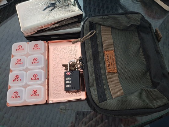 Auf einem Tisch liegt eine Pillendose und eine kleine Tasche. Die Pillendose ist aus rotgoldenem Metall, in den Deckel ist ein florales Muster gestanzt und sie ist aufgeklappt
Sie ist etwas größer als eine Spielkarte und enthält innen ein Plastik-Grid, in dem acht kleine herausnehmbare Behälter einsortiert sind, die jeweils mit einem Wochentag bedruckt sind.
