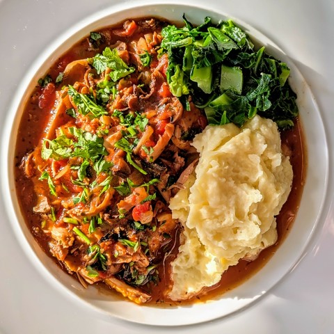 White bowl, chunky red stew with bits of chicken, onions and mushrooms visible, pile of mash, greens, parsley garnish
