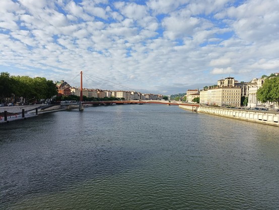 Photographie numérique couleur en format paysage.
Vue prise depuis un pont (Alphonse-Juin) avec une rivière (la Saône), une passerelle métallique en aval, des bâtiments et des arbres sur chaque rive, et un ciel bleu moutonné de nuages blancs.