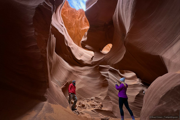 A slot canyon with smooth, curving walls in deep reddish-brown hues. Light from the open sky above filters down, illuminating the intricate patterns formed by erosion. Two people stand inside—one capturing a photo of the other—framing the natural beauty of the canyon. The scene highlights the dramatic contrast between shadowed rock formations and bright sunlight above, emphasizing the canyon's depth and textures.