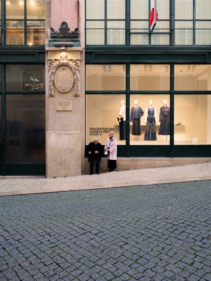 An elder couple rests in front of a clothes shop window. The man is sitting on the stone base of the window, while the woman is standing.