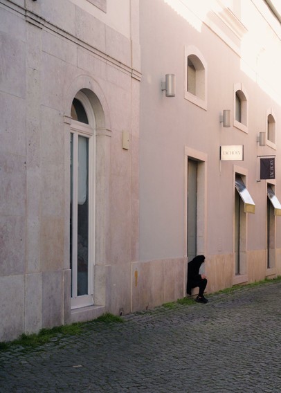 A yound woman wearing black clothes is sitting on a door sill while using her phone and smoking. Bright sunlight comes from above, but the alley is in the shade.