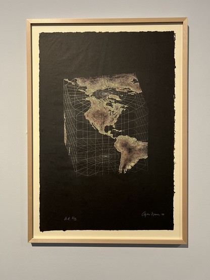 photo of a framed print showing a projection of the earth onto a cube-shaped grid. the continents are in brown colours and the oceans are empty and transparent and just show the silver grid lines of the cube structure. the map is printed onto black paper and signed by Agnes Denes in 1986 as print 2 of 15