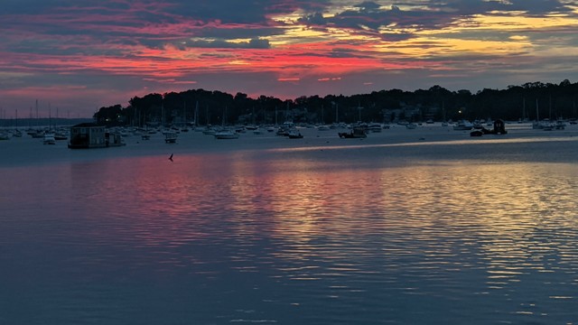 Historical Documentation in the form of a photograph of a rising sun over the ocean bay at the Historic Pioneer Village in Forest River Park, Salem, MA. The sky is filled with vibrant colors of pink, orange, and yellow, with the sun rising in the background behind the clouds. The water is calm and reflects the colors of the sky, creating a mirror-like effect. In the foreground, there are several boats and yachts docked in the harbor, with a few people walking along the shore. The harbor is surrounded by trees and buildings, and there is a small island in the distance. The overall mood of the image is peaceful and serene.