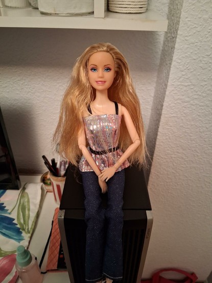 La muñeca sentada encima de mi PC con unos vaqueros, unos tacones plateados y un top brillante plateado y rosa con una cintura y tirantes negros