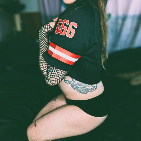 fox is kneeling on his black satin sheets wearing velvet black spanks and a cropped jersey that reads 666 with red and white stripes, he also has on fishnet gloves. his thighs are thick, and hairy and he has multiple tattoos and long red hair. one had is on his chest.