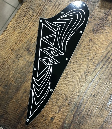 A black decorative pickguard featuring white geometric patterns, including triangular shapes and curved lines, laid on a wooden surface.