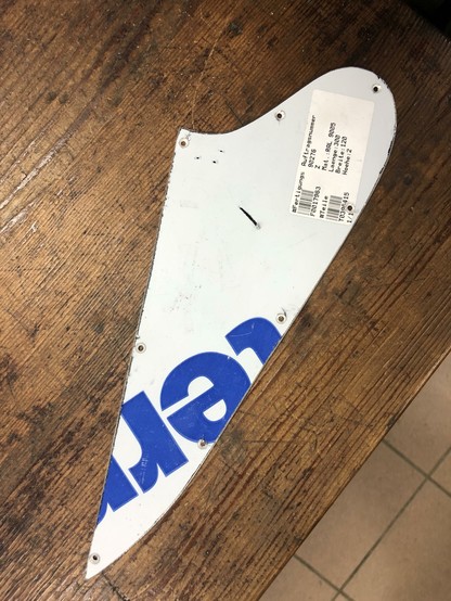A fragment of a white and blue sign or panel rests on a wooden surface, featuring text in blue. The panel has several holes along its edge, possibly for mounting, and a label with printed information attached to it.