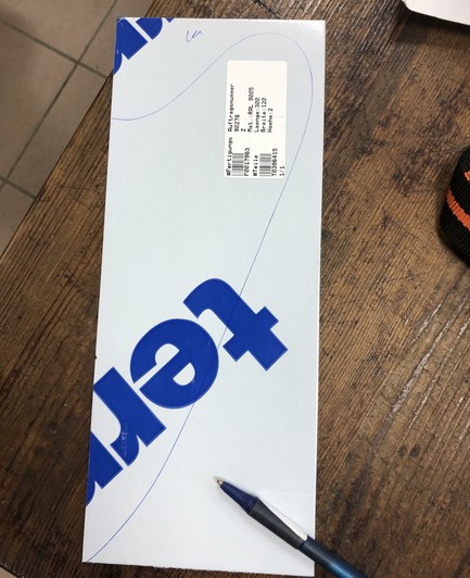 A foil covered metal sheet with a large blue logo and a label containing information. A blue pen is placed next to it, on a wooden table surface. The lines of a pickguard are marked on the foil.