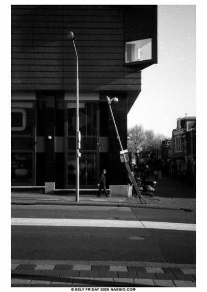 Black and white photograph looking across a street on a slight slope to the right at a building with a window on the corner. On the sidewalk in front is a lamp post and a surveillance camera lamppost that looks like it has been hit by a car as it is bent, leaning almost 45˚. Between the two posts is a person walking left.