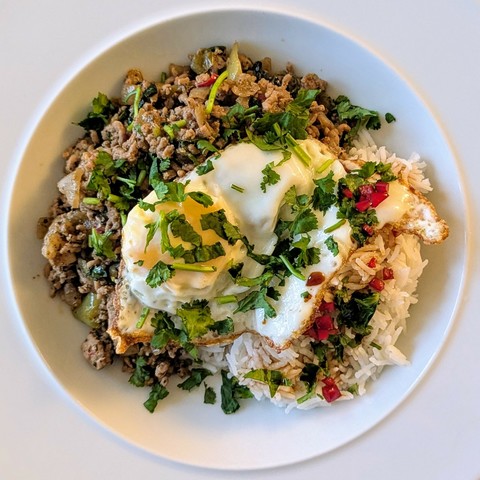 White bowl, pork mince with bits of green and red, pile of rice with a little dark sauce dotted with chilli, fried egg over both, chopped coriander