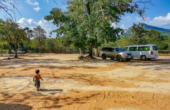 Photo shows a large sand covered area with some trees and cars parked in their shadow. It is a sunny day with some blue sky visible in the distance. Running into the sandy area is a 2-year-old boy dressed in shorts only. There is obvious playtime and breaktime in the sun.