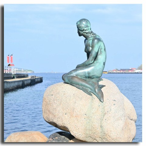 Die Kleine Meerjungfrau in Kopenhagen bei blauem Himmel und Sonnenschein von ihrer linken Seite aus gesehen. Meerjungfrau und Bildbetrachter kucken in dieselbe Richtung zur offenen Ostsee bzw. zum Öresund.