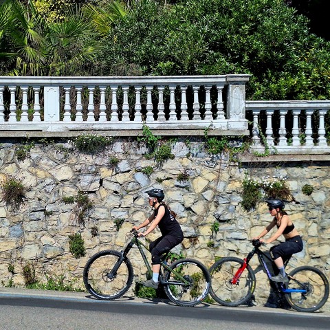 Picture of a stone masonry retaining wall, topped with a balustrade featuring small stone columns. Two people on bikes are riding uphill in front of the wall.