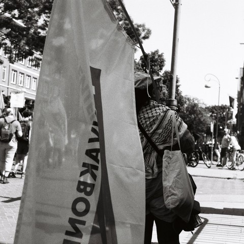 B&W image worker unions demonstrating on May first union member with AKKU flag observing bystanders and march, student workers union, bystanders unsharp in background