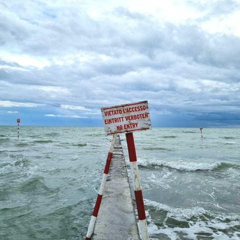 A jetty juts out into the choppy sea under a cloudy sky. Two wooden poles marked red and white are mounted crosswise on the jetty holding the sign "VIETATO L'ACCESSO. EINTRITT VERBOTEN. NO ENTRY".