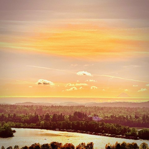 A radiant orange sky shines brightly on the trees, hills, mountains, and single white building below whose features somewhat resemble a cartoon face with furrowed brows, all on a partly cloudy morning.