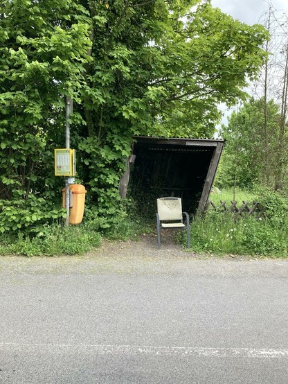 Bushaltestelle von gegenüber fotografiert, über die grau asphaltierte Straße hinweg. Darum ist diese im Vordergrund zu sehen. Dann links der Pfahl an dem Papierkorb (orangefarben) und der Busfahrplan im gelben Rahmen befestigt sind. In der Mitte steht eine hölzerne windschiefe Bude mit einem grauen Gartenstuhl mit hellgrauem Plastikgeflecht, welches oben an der Lehne defekt ist. Rundherum ist es grün, links mehr Bäume, rechts Wiese.