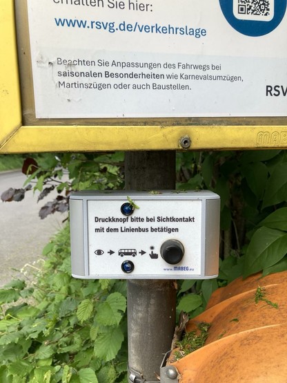 Hier gibt es ein Ding zu sehen, auf dem steht; Druckknopf bitte bei Sichtkontakt mit dem Linienbus betätigen. Darunter ist ein Auge abgebildet, dann folgt ein Pfeil, dann ein Linienbussymbol, dann eine schwarze Hand deren Zeigefinger auf den Druckknopf tippt. Daneben befindet sich dann ein dicker schwarzer echter Druckknopf. Das Ding ist unter dem Busfahrplan am Pfahl montiert.