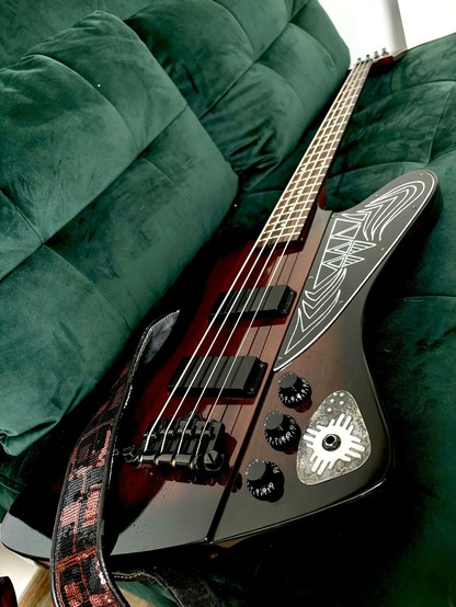 An electric bass guitar rests on a green velvet couch. The guitar features a unique design on its body and has several control knobs. The cozy couch background contrasts with the instrument's glossy finish.