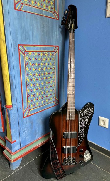 An electric bass guitar with a unique body shape and black and brown finish. The pickguard is black engraved with curvy and triangular shapes, the jack has a silver triangular shaped cover of vintage finished silver with four golden stars. The guitar positioned against a colorful, intricately designed wooden cabinet with a blue background.