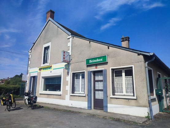 vue extérieure d'une maison avec 2 vélos stationnés devant. ciel bleu. maison en 2 partie : à gauche une épicerie avec une devanture Proxy , à droite un bar avec un panneau d'une marque de bière au dessus de la porte d'entrée avec des vieux rideaux à l'ancienne