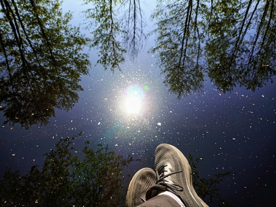 Meine Füße baumeln über Wasser. Im Wasser spiegeln sich die Sonne sowie die Bäume am gegenüberliegenden Ufer.