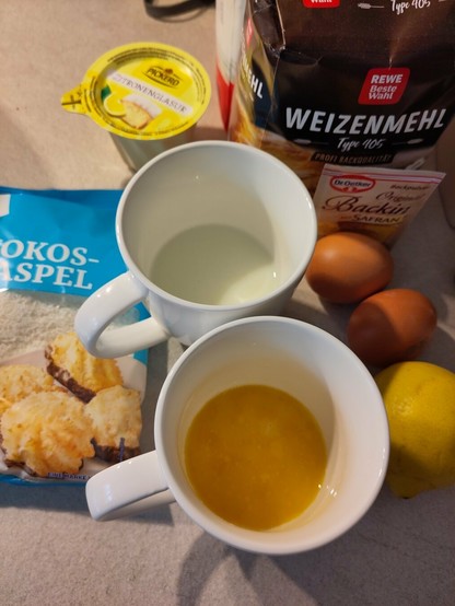 Die Zutaten für den Kuchen, darunter flüssige Margarine, Eier, Zitrone, Kokosflocken, Mehl und Zucker.