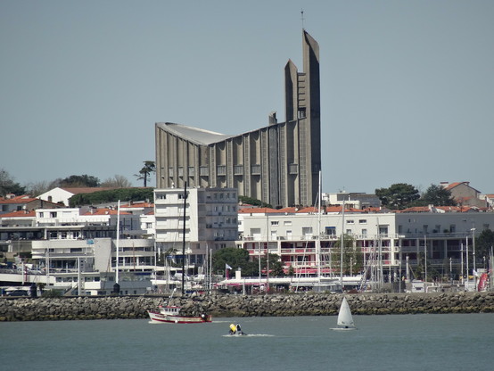 Blick vom Meer aus auf Häuser an der bebauten Küste. Im Vordergrund noch auf dem Meer ist ein kleines Boot, ein Surfer und ein Motorboot zu sehen. Hinter der Häuserfront steht eine Kirche aus Beton im Stile von Notre Dame.