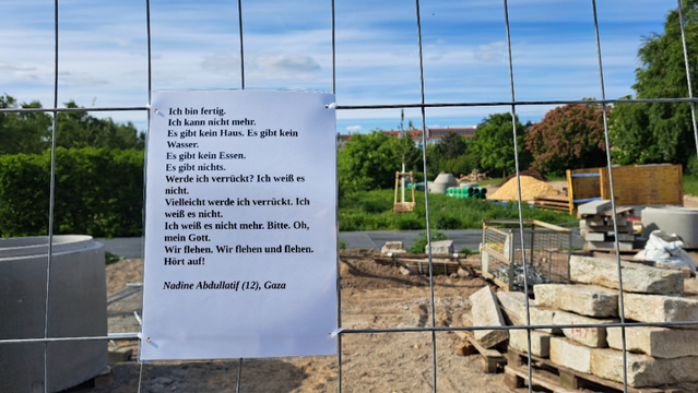 An einem Bauzaun aufgehängtes, laminiertes A4 Blatt mit folgendem Text: 
"Ich bin fertig.
Ich kann nicht mehr.
Es gibt kein Haus. Es gibt kein Wasser.
Es gibt kein Essen.
Es gibt nichts.
Werde ich verrückt? Ich weiß es nicht.
Vielleicht werde ich verrückt. Ich weiß es nicht.
Ich weiß es nicht mehr. Bitte. Oh, mein Gott.
Wir flehen. Wir flehen und flehen.
Hört auf!

Nadine Abdullatif (12), Gaza"

im Hintergrund Bäume und blauer Himmel, direkt hinter dem Bauzaun Erde und Baustellengegenstände