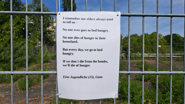 Laminated A4 sheet with the following text hung on a construction fence. In the background, trees and blue sky, earth and construction site objects directly behind the fence

"I remember our elders always used to tell us - 
No one ever goes to bed hungry.
No one dies of hunger in their  homeland. 
But every day, we go to bed hungry. 
If we don’t die from the bombs, we'll die of hunger.

Eine Jugendiiche (13), Gaza"