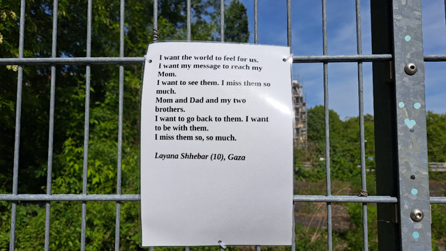 Laminated A4 sheet with the following text hung on a construction fence. In the background, trees and blue sky

"I want the world o feel for us
I want my message to reach my Mom. 
I want to see them. I miss them so much.
Mom and Dad and my two brothers. I want to go back to them. I want to be with them. I miss them so, so much."
 
Layana Shhebar (10), Gaza