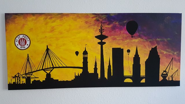 Bildbeschreibung von KI:
Dieses Foto zeigt ein gemaltes Bild mit der Skyline von Hamburg in stilisierter Silhouette. Die Gebäude und Wahrzeichen – wie der Fernsehturm (Heinrich-Hertz-Turm), der Michel (St. Michaelis Kirche), die Elbphilharmonie und verschiedene Hafenkräne – sind in Schwarz gehalten.

Im Hintergrund strahlt ein farbenfroher Sonnenuntergang in Gelb-, Orange-, Rot- und Violetttönen. Außerdem sind einige Heißluftballons zu sehen, die dem Bild eine verspielte Note verleihen. Links oben ist das Logo des Fußballvereins FC St. Pauli mit der Jahreszahl 1910 prominent zu erkennen – ein klarer Hinweis auf die Verbundenheit des Künstlers oder der Besitzerin/des Besitzers mit dem Stadtteil oder dem Verein.