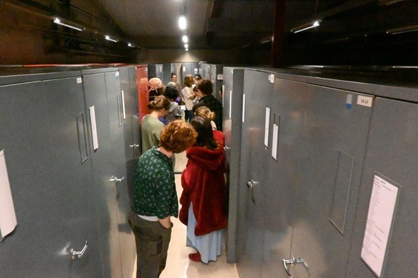 A photo of students gathered between a row of tall cabinets. At the back botanist Sue Gibbs has a cabinet open and is talking about some grass specimens.

My full-res photo is on Flickr at https://www.flickr.com/photos/mollivan_jon/54505210018/