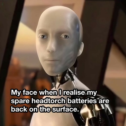 A humanoid robot from "I, Robot" with a smooth, featureless face and a shiny metallic body. Its expression appears humorously deadpan with a tinge of exasperation. The robot's head is bald, and its eyes are blue, giving it a somewhat lifelike, yet robotic, appearance. The background suggests a futuristic or artificial setting, with soft lighting and dark, angular shapes. In the lower part of the image, there's a caption reading, "My face when I realise my spare headtorch batteries are back on the surface," adding a humorous context to the robot's expression.