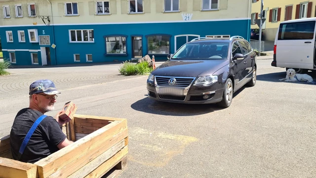 halb-petrolfarbenes Gebäude Krone Randegg unterer Teil gestrichen oberer noch alt-gelb-grün-dreckig auf der anderen Straßenseite im Vordergrund sitzt ein Mann mit Schirmmütze in einem palettengroßen Pflanzkübel aus Altholz, davor zwei Autos vor dem Bussel liegt ein weißer Golden Retriever
