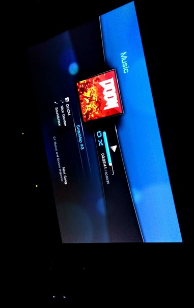 The photograph composed in total pitch black darkness shows a screen displaying a music player interface. The background is predominantly blue with a darker blue section on the left. The word "Music" is displayed in white text on the right side. In the center, there is a red and orange graphic with the text "DOOM" in white, which is the album cover for the track being played. Below the album cover, the track title "DOOM" and the artist "Mick Gordon" are shown. The playback controls are visible, including a progress bar indicating the current time (00:24.1) out of a total duration (00:03.35). The playback speed is set to 0x. The next song is listed as "01. Mystic and Severe (inground)" from "SnapMap #3." The interface also includes navigation buttons on the left side. The screen is set against a dark background, emphasizing the interface elements.

 Ovis2-8B

🌱 Energy used: 0.233 Wh
