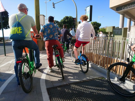 Numeroso grupo de ciclistas* en un carril bici sobre acera peatonal esperando la apertura del semáforo. Están montados en distintos tipos de bicicletas de alquiler, de color verde o azul. Las bicicletas azules son públicas, gracias al sistema de Bicicoruña, mientras que las verdes parece que son de alguna empresa de bicicletas que las alquila.

*Entre 8 y 10, sin contar conmigo.
