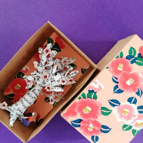 Un arbre en papier blanc dans une petite boîte qui peut se refermer (comme une grande boîte d'allumette). L'arbre est recouvert de texte en français et japonais, imprimé en noir (on y aperçoit notamment le mot "cerisier"). On y voit également quelques fleurs de cerisiers roses au niveau des feuilles. Une femme vue de côté se tient sous l'arbre en tailleur, face à ce dernier. Le fond et le dessus de la boîte est rose clair, avec des motifs de fleurs de cerisiers roses et de feuilles vertes.
