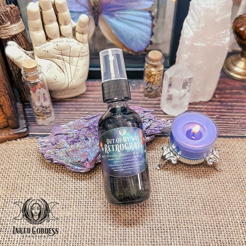 A small smoked glass spray bottle of "Out of My Way, Retrograde Mist" is propped on a piece of chalcopyrite on a burlap-covered surface. It's surrounded by crystals with a tealight for scale. The label on the bottle includes the name of the product, its metaphysical properties, and its ingredients. This is an exclusive product from Inked Goddess Creations.